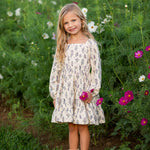 A young girl stands smiling in a field of wildflowers, holding a pink flower, her Gracie Long Sleeve Floral Square Neck Tiered Dress from Mabel and Honey beautifully echoing the colorful blooms and lush green grass that surround her.