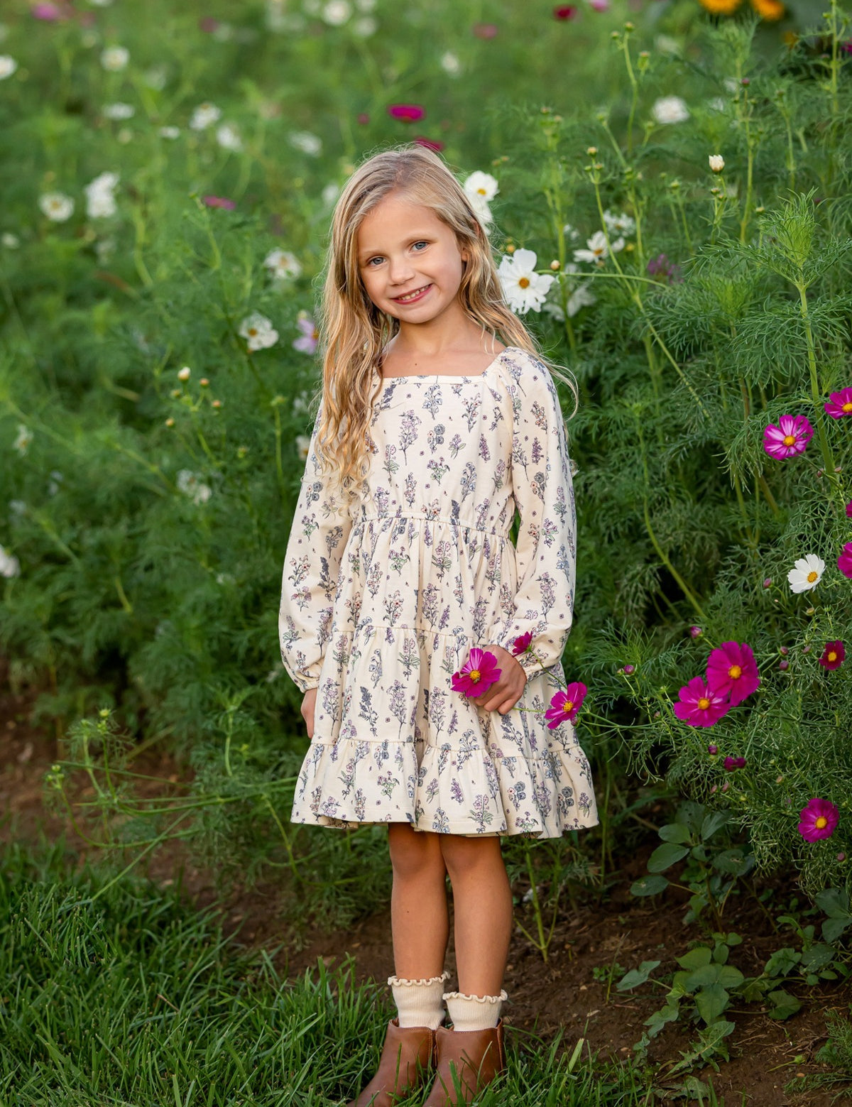 A young girl stands smiling in a field of wildflowers, holding a pink flower, her Gracie Long Sleeve Floral Square Neck Tiered Dress from Mabel and Honey beautifully echoing the colorful blooms and lush green grass that surround her.