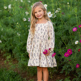 A young girl stands smiling in a field of wildflowers, holding a pink flower, her Gracie Long Sleeve Floral Square Neck Tiered Dress from Mabel and Honey beautifully echoing the colorful blooms and lush green grass that surround her.
