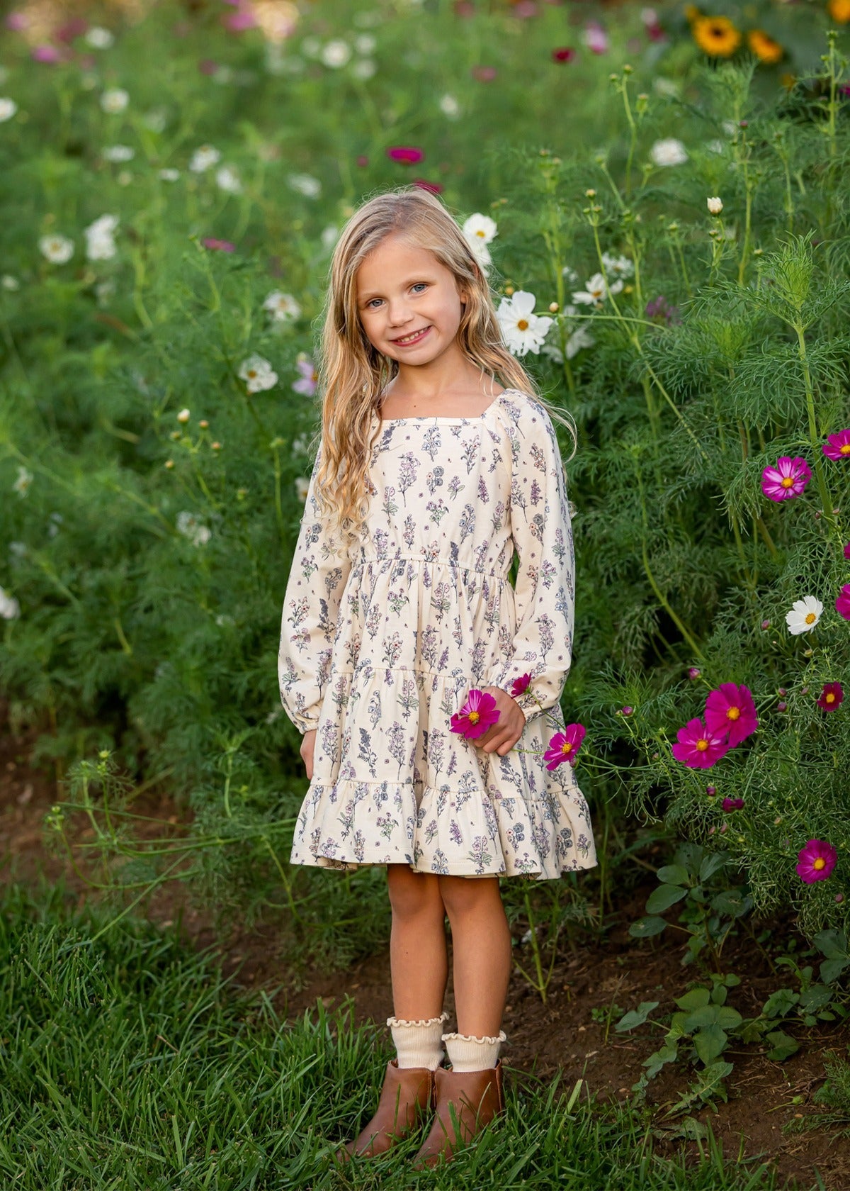 A young girl stands smiling in a field of wildflowers, holding a pink flower, her Gracie Long Sleeve Floral Square Neck Tiered Dress from Mabel and Honey beautifully echoing the colorful blooms and lush green grass that surround her.