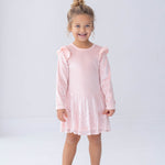 A young girl with curly hair, dressed in the Isobella and Chloe Blushing Velvet Long Sleeve Ruffle Shoulder Sequin Skirt Dress and silver shoes, stands smiling against a plain white background—perfect for festive holiday parties.