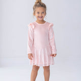 A young girl with curly hair, dressed in the Isobella and Chloe Blushing Velvet Long Sleeve Ruffle Shoulder Sequin Skirt Dress and silver shoes, stands smiling against a plain white background—perfect for festive holiday parties.