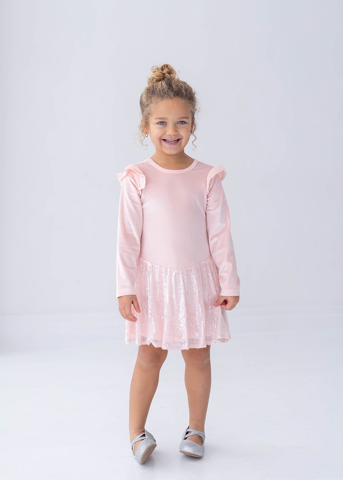 A young girl with curly hair, dressed in the Isobella and Chloe Blushing Velvet Long Sleeve Ruffle Shoulder Sequin Skirt Dress and silver shoes, stands smiling against a plain white background—perfect for festive holiday parties.