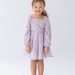 A young child with curly hair is standing and smiling against a plain white background, wearing the Isobella and Chloe Sugarplum Sparkle Long Sleeve Floral Sequin Square Neck Tiered Dress paired with silver shoes.