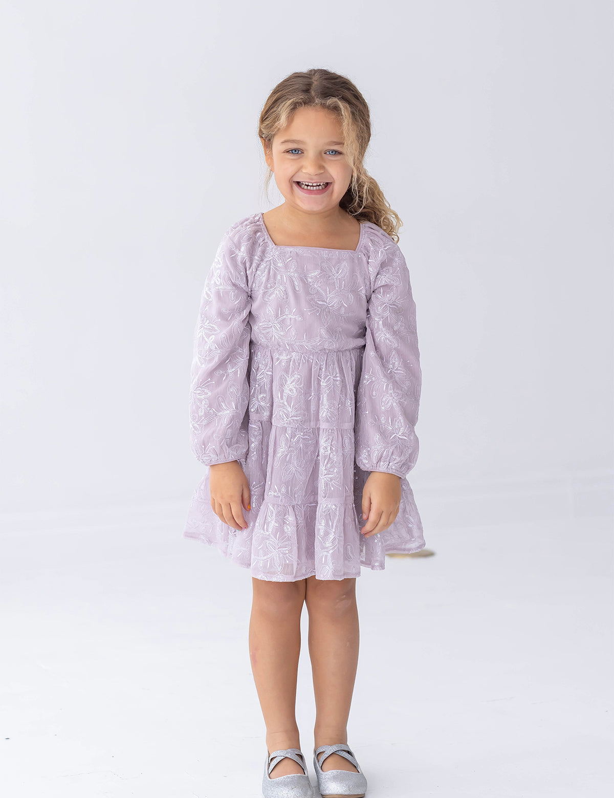 A young child with curly hair is standing and smiling against a plain white background, wearing the Isobella and Chloe Sugarplum Sparkle Long Sleeve Floral Sequin Square Neck Tiered Dress paired with silver shoes.