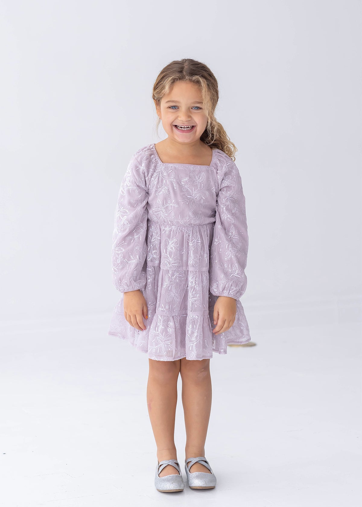 A young child with curly hair is standing and smiling against a plain white background, wearing the Isobella and Chloe Sugarplum Sparkle Long Sleeve Floral Sequin Square Neck Tiered Dress paired with silver shoes.