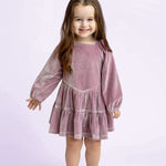 A young girl with long brown hair smiles while wearing the Isobella and Chloe Sugarplum Sparkle Long Sleeve Raglan Tiered Velvet Dress, paired with sparkly shoes. She stands against a light purple background.