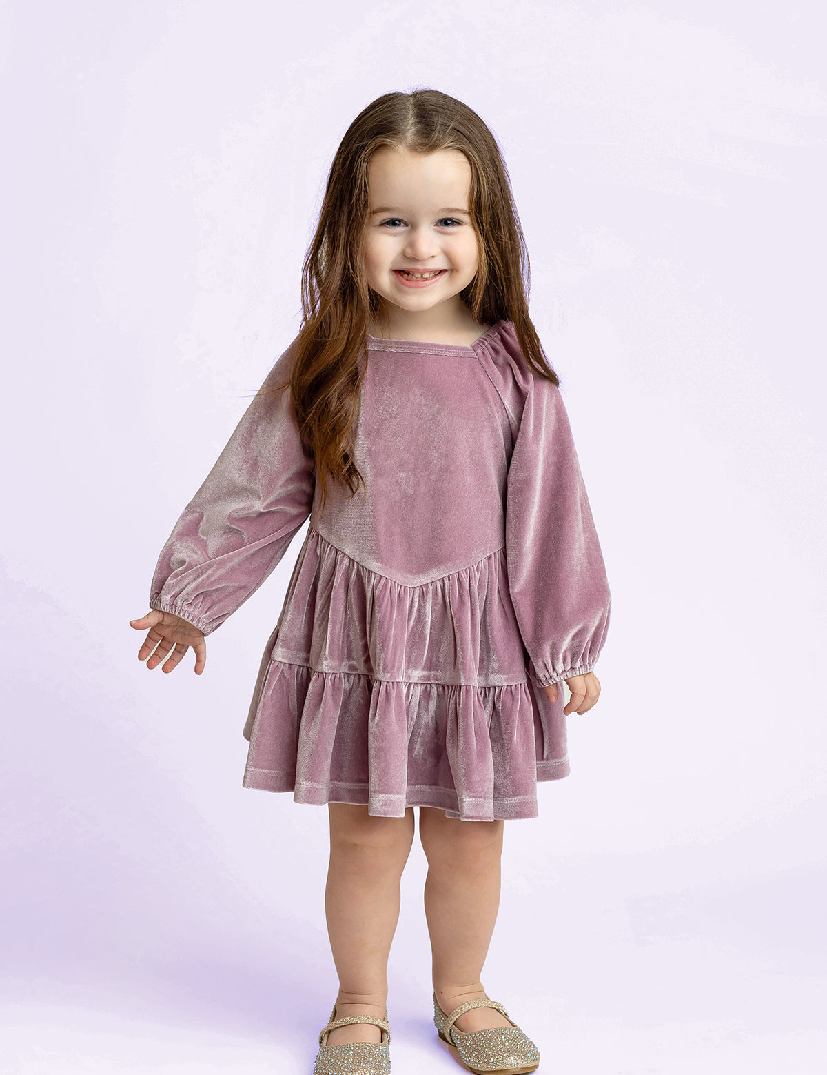 A young girl with long brown hair smiles while wearing the Isobella and Chloe Sugarplum Sparkle Long Sleeve Raglan Tiered Velvet Dress, paired with sparkly shoes. She stands against a light purple background.