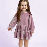 A young girl with long brown hair smiles while wearing the Isobella and Chloe Sugarplum Sparkle Long Sleeve Raglan Tiered Velvet Dress, paired with sparkly shoes. She stands against a light purple background.