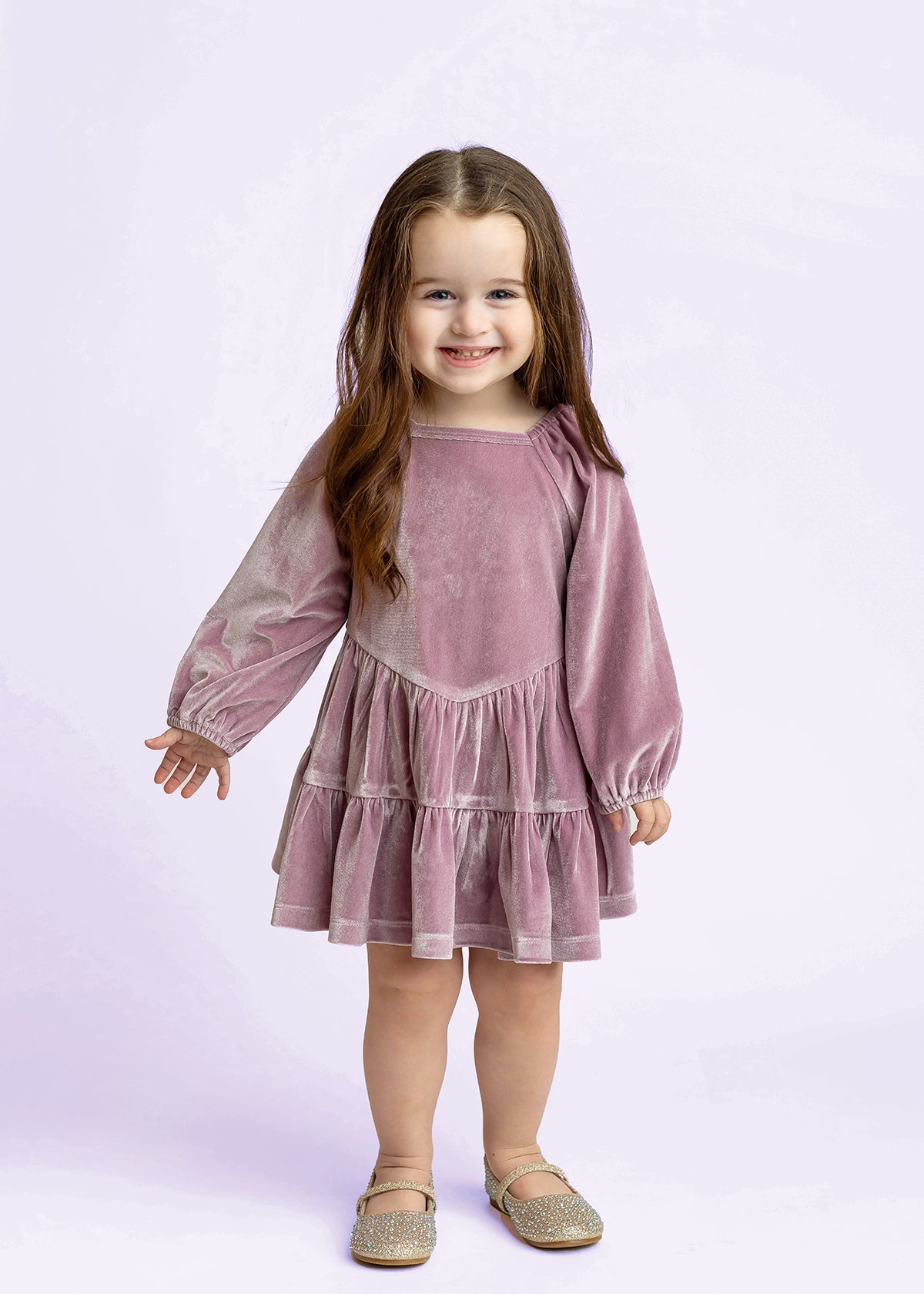 A young girl with long brown hair smiles while wearing the Isobella and Chloe Sugarplum Sparkle Long Sleeve Raglan Tiered Velvet Dress, paired with sparkly shoes. She stands against a light purple background.