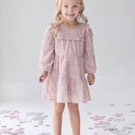 A young girl with long blonde hair wears the Isobella and Chloe Dusty Rose Long Sleeve Floral Sequin Ruffle Yoke Tiered Dress, paired with gray shoes. She stands smiling on a white floor decorated with scattered pastel and silver stars.