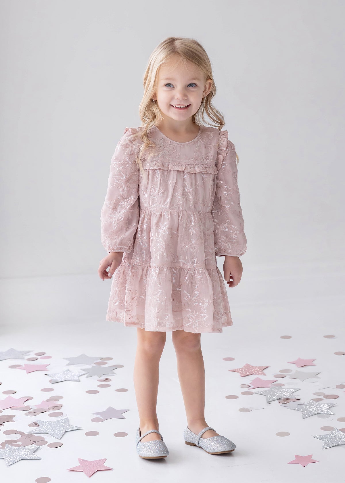 A young girl with long blonde hair wears the Isobella and Chloe Dusty Rose Long Sleeve Floral Sequin Ruffle Yoke Tiered Dress, paired with gray shoes. She stands smiling on a white floor decorated with scattered pastel and silver stars.