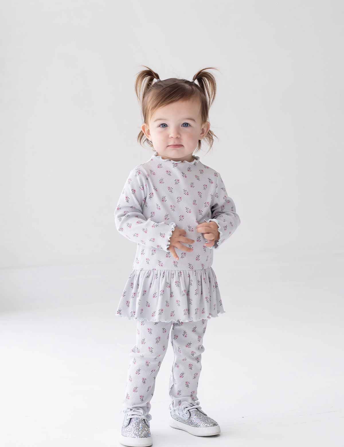 A toddler with pigtails stands against a white background, wearing the Mabel and Honey Petite Petal Long Sleeve Floral Ribbed Peplum Top and Legging Set paired with sparkly silver shoes—a charming addition to any girl’s clothing collection.