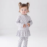 A toddler with pigtails stands against a white background, wearing the Mabel and Honey Petite Petal Long Sleeve Floral Ribbed Peplum Top and Legging Set paired with sparkly silver shoes—a charming addition to any girl’s clothing collection.