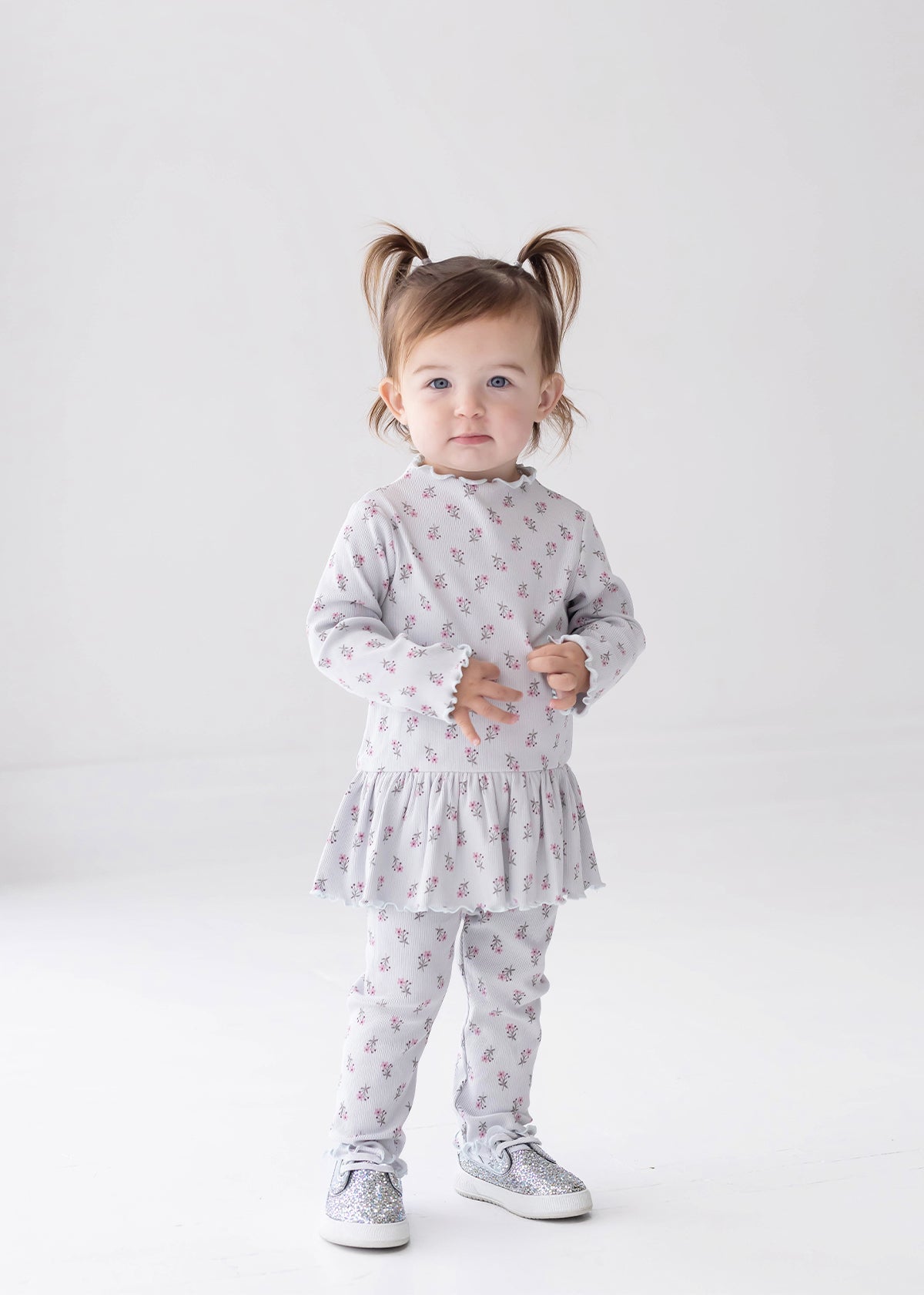 A toddler with pigtails stands against a white background, wearing the Mabel and Honey Petite Petal Long Sleeve Floral Ribbed Peplum Top and Legging Set paired with sparkly silver shoes—a charming addition to any girl’s clothing collection.