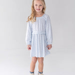 A young girl with long blonde hair is wearing the Mabel and Honey Luna Bella Long Sleeve Star Print Ruffle Trim A Line Dress, paired with white boots, as she stands smiling in a bright, minimalist studio with a white background.