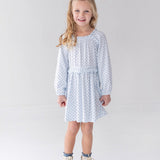 A young girl with long blonde hair is wearing the Mabel and Honey Luna Bella Long Sleeve Star Print Ruffle Trim A Line Dress, paired with white boots, as she stands smiling in a bright, minimalist studio with a white background.
