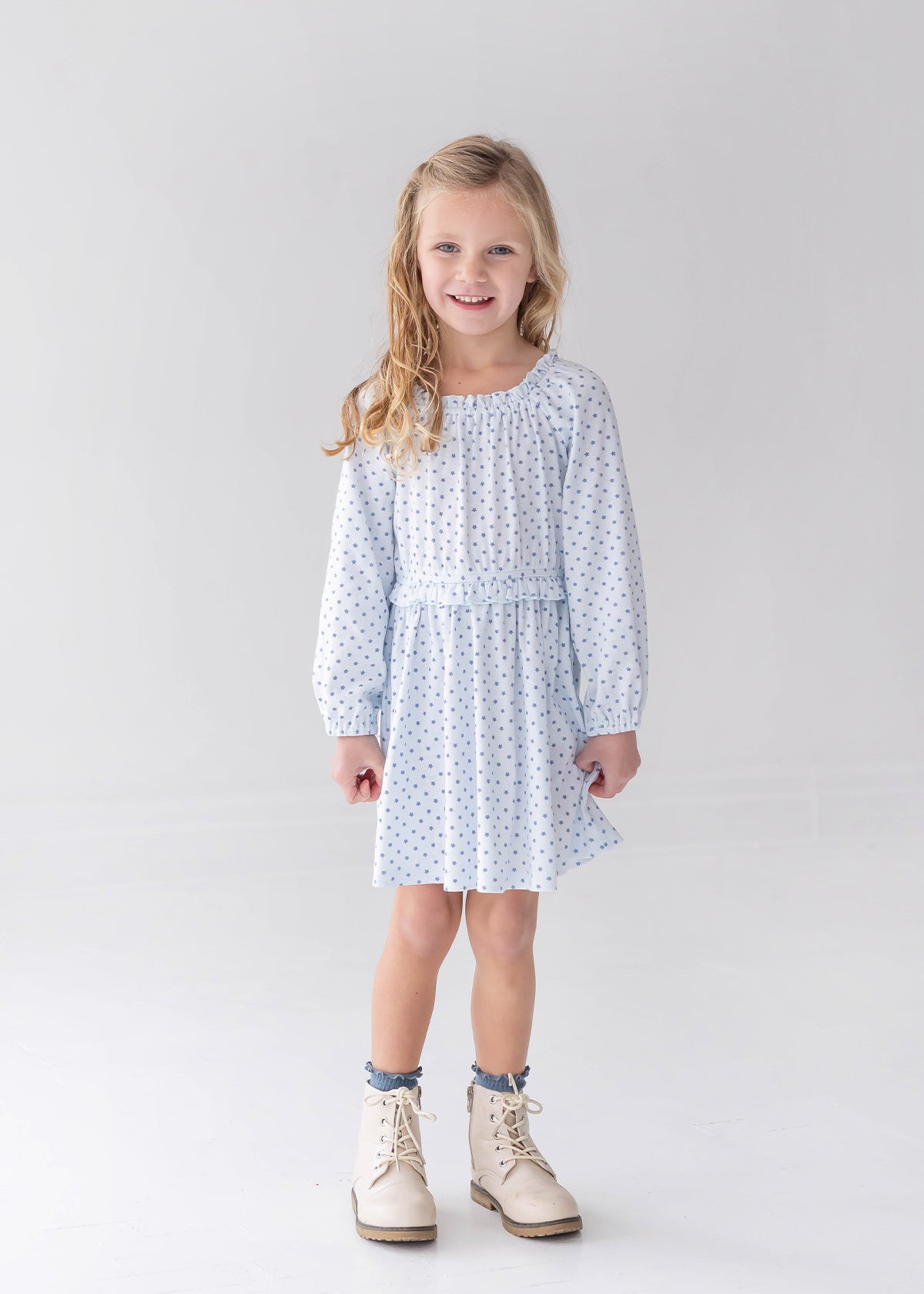 A young girl with long blonde hair is wearing the Mabel and Honey Luna Bella Long Sleeve Star Print Ruffle Trim A Line Dress, paired with white boots, as she stands smiling in a bright, minimalist studio with a white background.