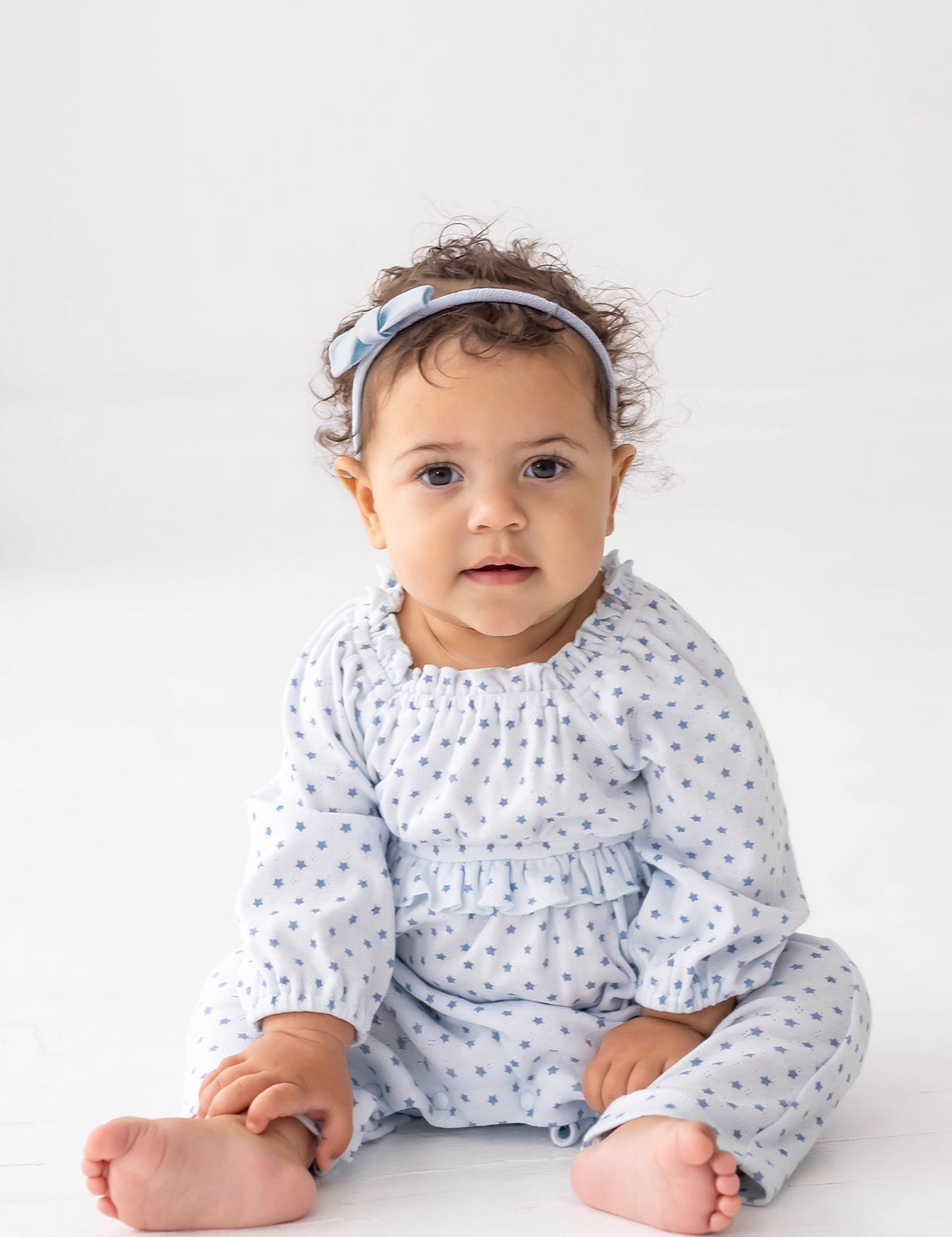 A baby with curly hair, barefoot and wearing the Mabel and Honey Luna Bella Long Sleeve Star Print Ruffle Trim Romper—a soft cotton blend blue outfit adorned with stars—paired with a matching blue headband, sits on a white floor against a plain white background.
