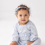 A baby with curly hair, barefoot and wearing the Mabel and Honey Luna Bella Long Sleeve Star Print Ruffle Trim Romper—a soft cotton blend blue outfit adorned with stars—paired with a matching blue headband, sits on a white floor against a plain white background.