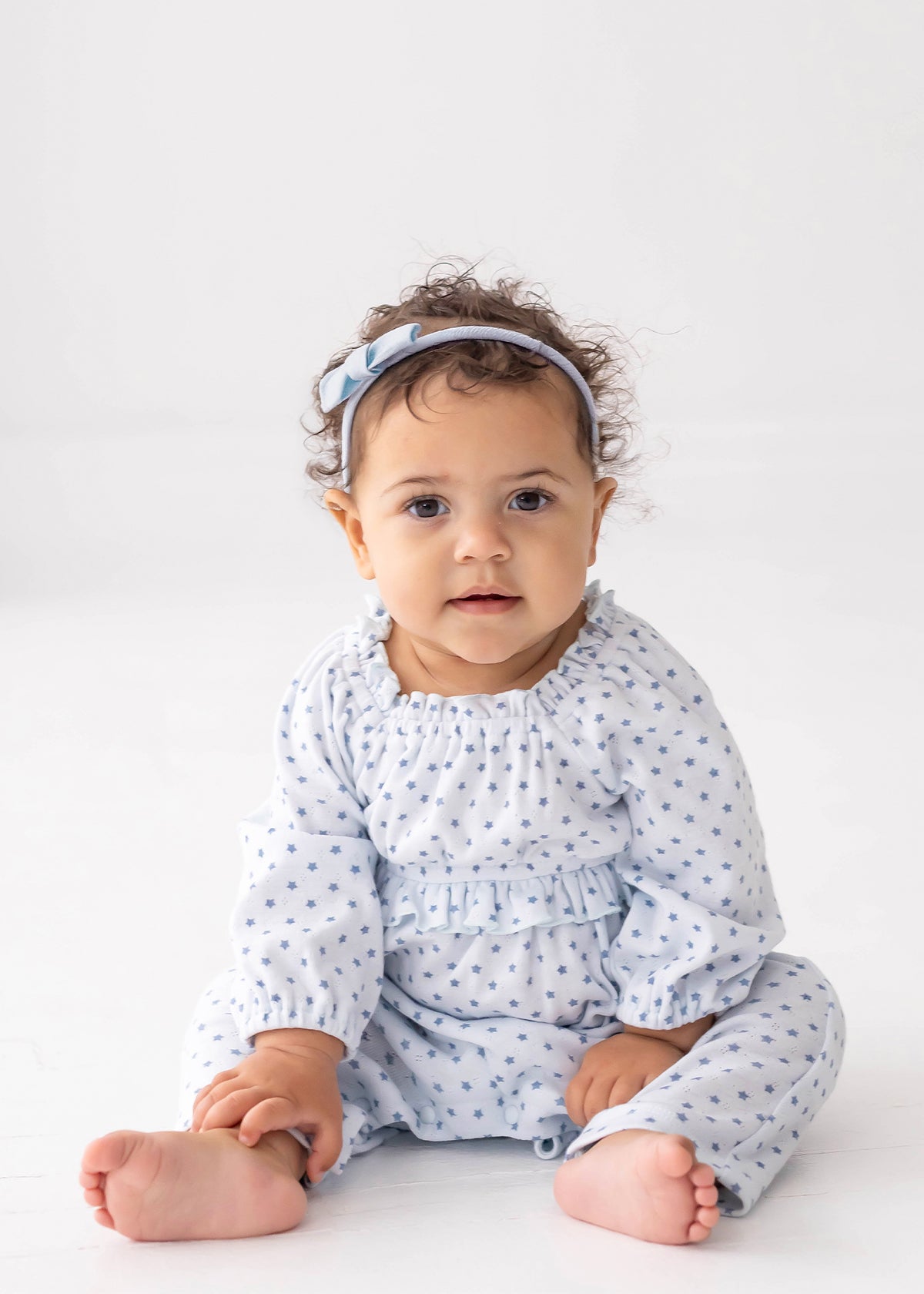 A baby with curly hair, barefoot and wearing the Mabel and Honey Luna Bella Long Sleeve Star Print Ruffle Trim Romper—a soft cotton blend blue outfit adorned with stars—paired with a matching blue headband, sits on a white floor against a plain white background.