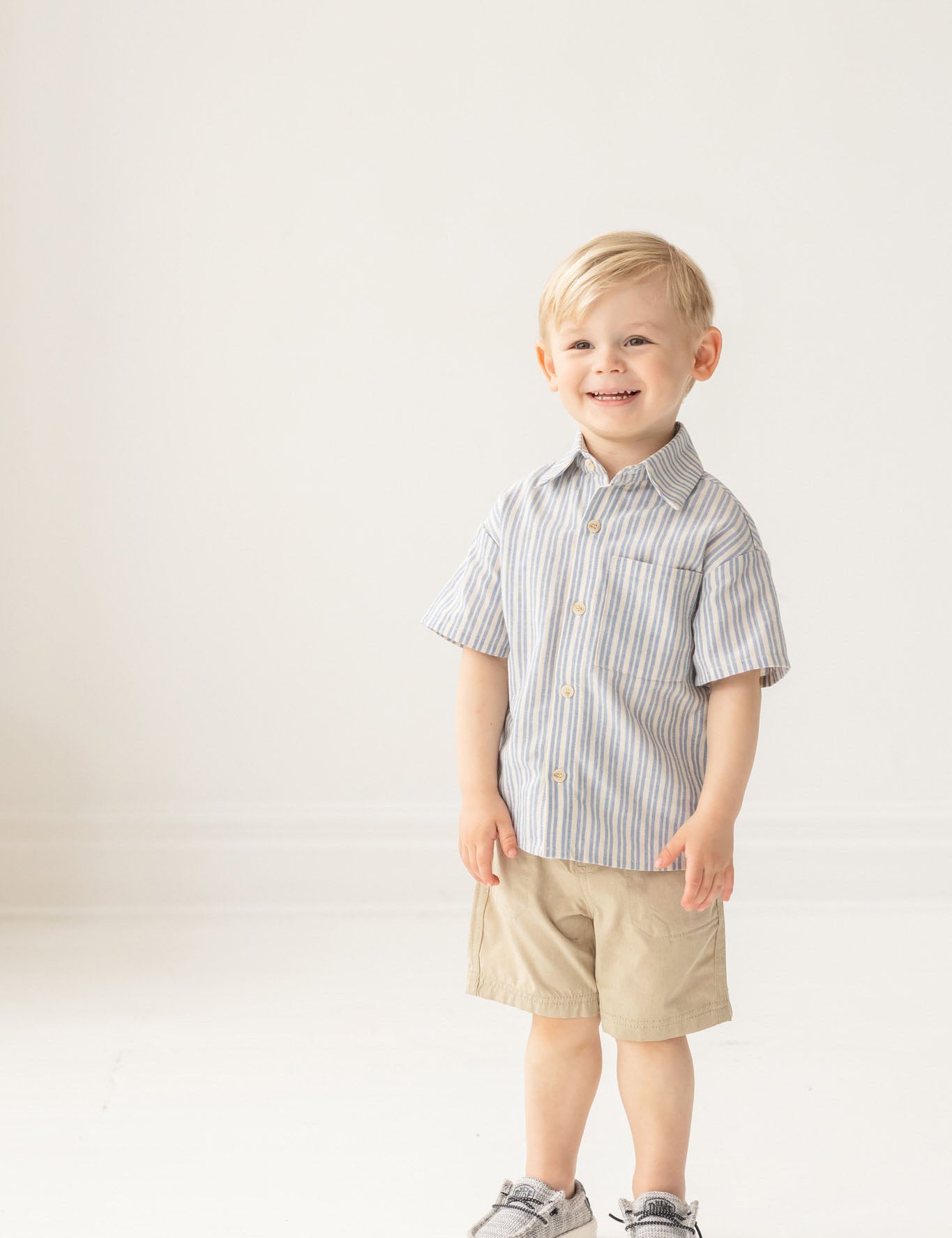 A young boy with blond hair smiles against a plain white background, wearing the Mabel and Honey Breezy Blue Skies Top—a striped cotton blend short-sleeve shirt—paired with tan shorts and gray shoes.