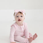 A baby sits barefoot on a white floor, wearing the Mabel and Honey Amelia Rose Long Sleeve Solid Romper with ruffles and a matching pink bow headband, looking up with an open mouth.