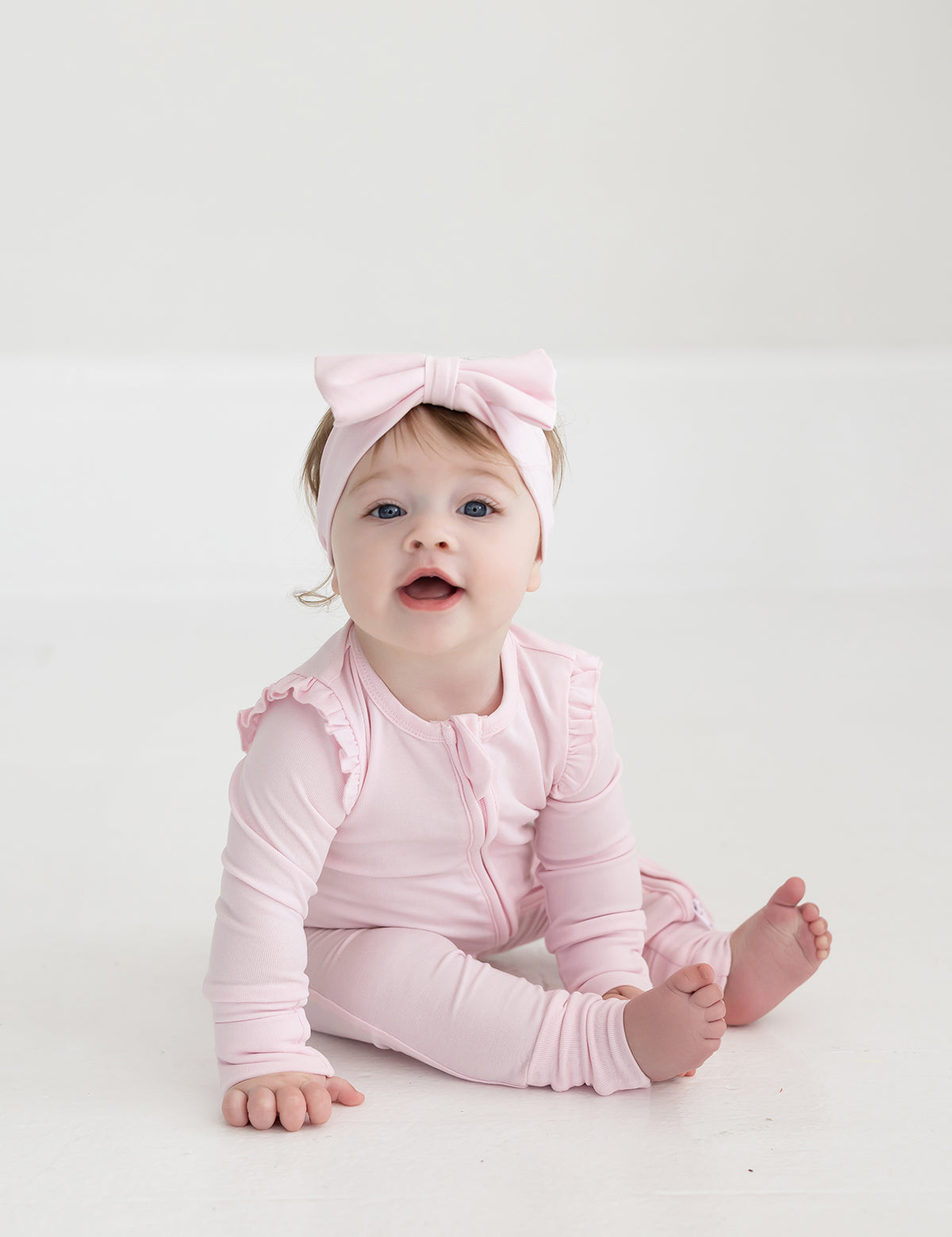 A baby sits barefoot on a white floor, wearing the Mabel and Honey Amelia Rose Long Sleeve Solid Romper with ruffles and a matching pink bow headband, looking up with an open mouth.
