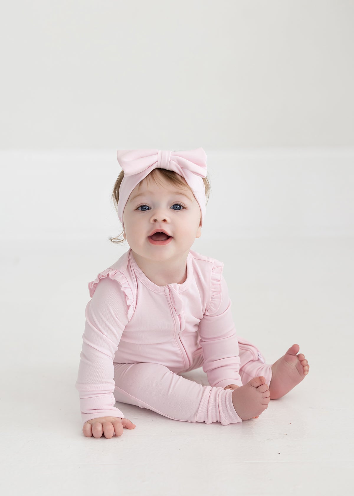 A baby sits barefoot on a white floor, wearing the Mabel and Honey Amelia Rose Long Sleeve Solid Romper with ruffles and a matching pink bow headband, looking up with an open mouth.