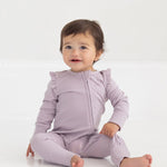 A baby with short brown hair sits on a white floor, smiling slightly and wearing the Mabel and Honey Soft Meadow Long Sleeve Solid Convertible Romper with Matching Headband, which features ruffled shoulders and a convenient two-way front zipper.