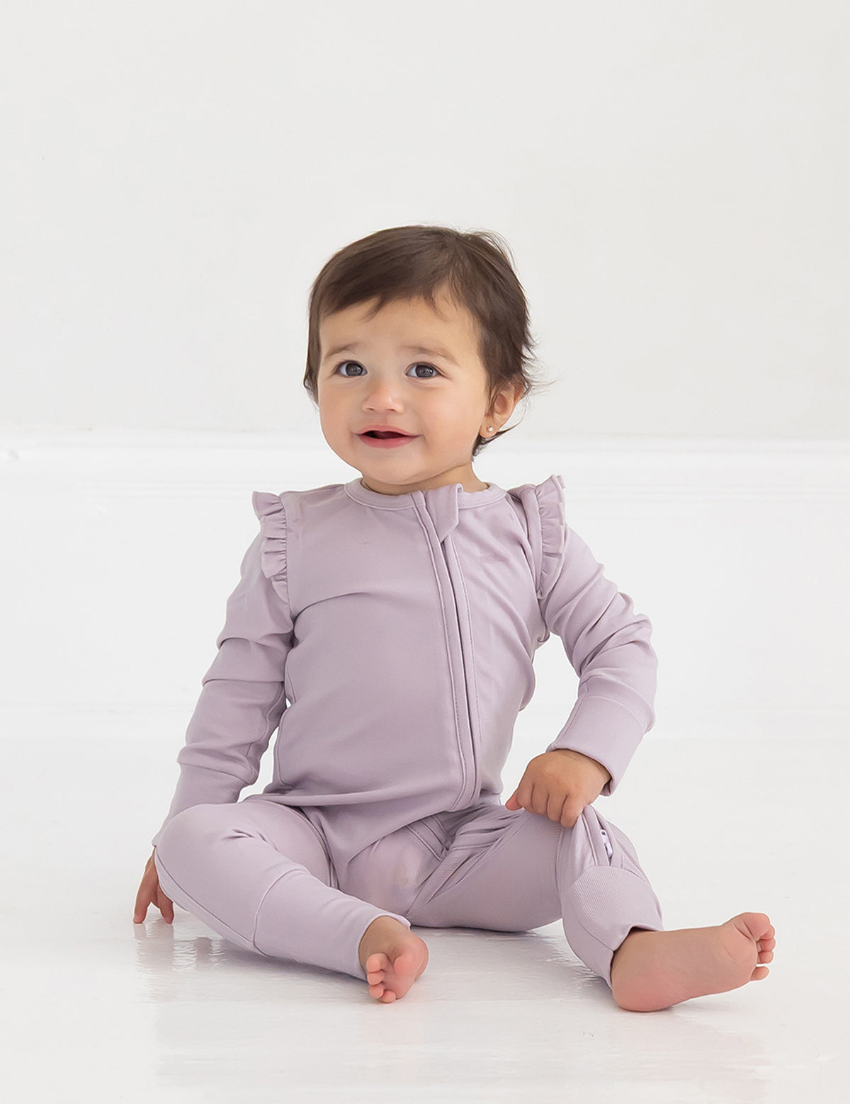 A baby with short brown hair sits on a white floor, smiling slightly and wearing the Mabel and Honey Soft Meadow Long Sleeve Solid Convertible Romper with Matching Headband, which features ruffled shoulders and a convenient two-way front zipper.