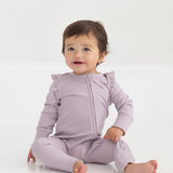 A baby with short brown hair sits on a white floor, smiling slightly and wearing the Mabel and Honey Soft Meadow Long Sleeve Solid Convertible Romper with Matching Headband, which features ruffled shoulders and a convenient two-way front zipper.