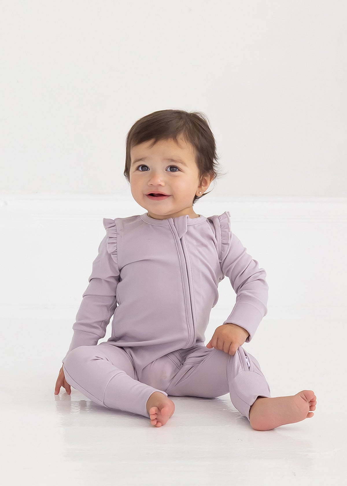 A baby with short brown hair sits on a white floor, smiling slightly and wearing the Mabel and Honey Soft Meadow Long Sleeve Solid Convertible Romper with Matching Headband, which features ruffled shoulders and a convenient two-way front zipper.