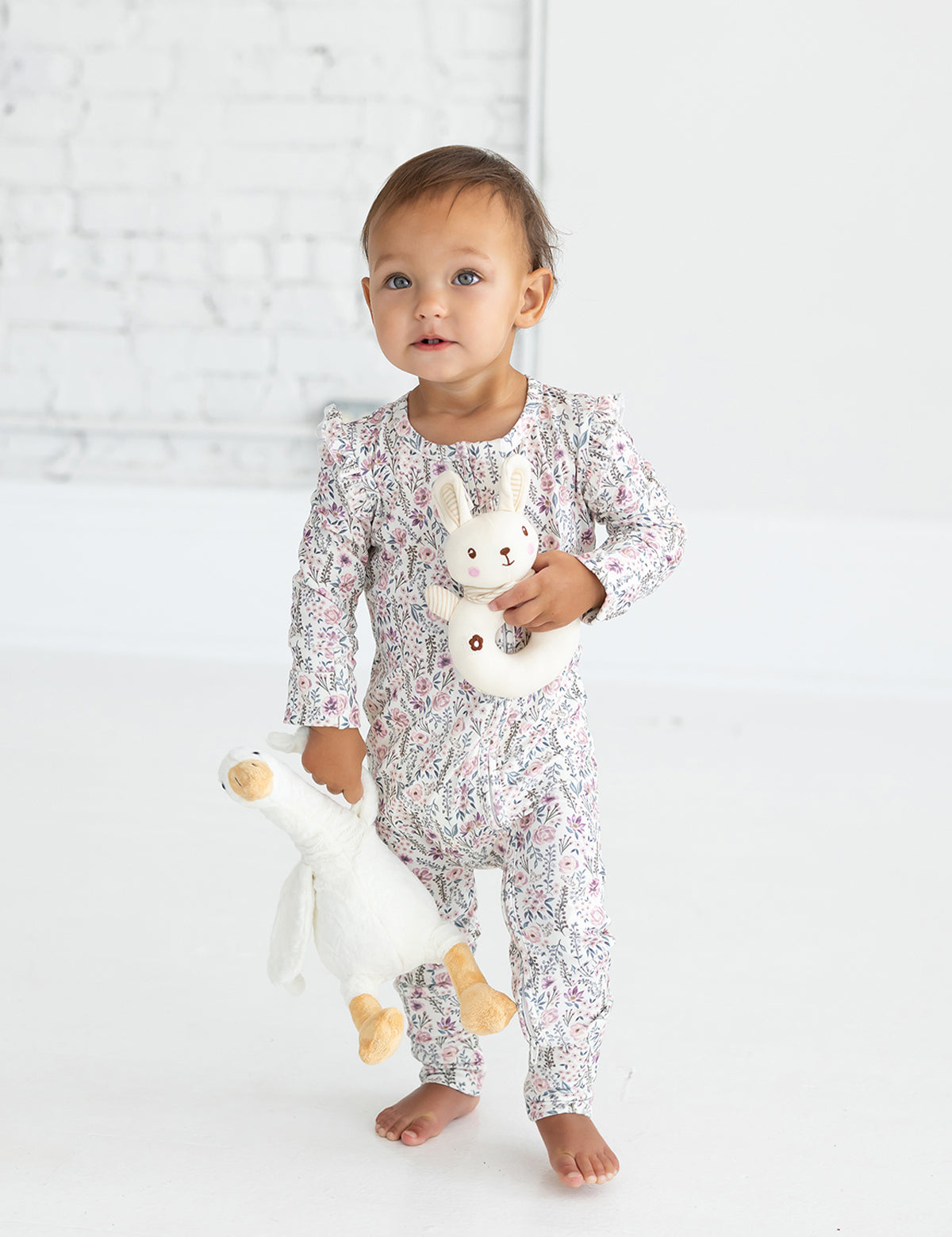 A toddler wearing the Mabel and Honey Whimsical Wildflower Long Sleeve Floral Print romper with a matching headband stands barefoot on a white floor, holding a plush white goose in one hand and a bunny rattle in the other, with a white brick wall in the background.
