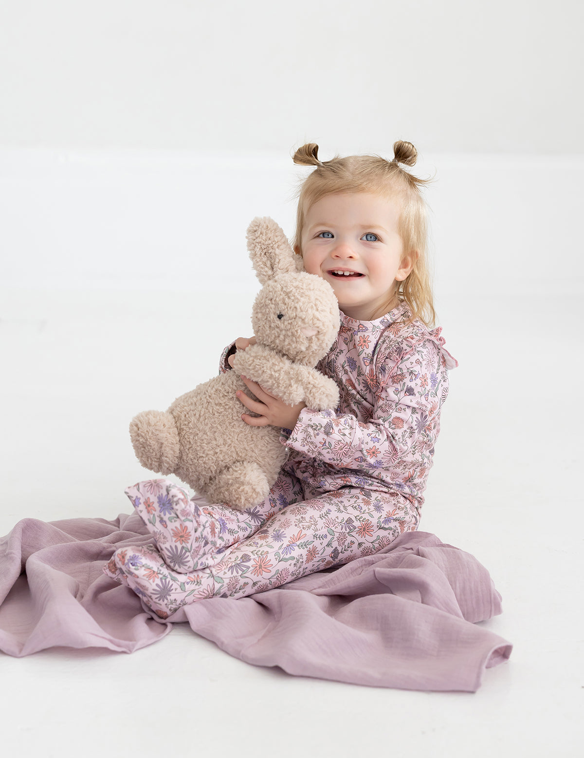 A young child with blonde pigtails beams as she sits on a lilac blanket in a bright, white room, hugging a plush bunny and wearing the Mabel and Honey Soft Meadow Long Sleeve Floral Print with Matching Headband.