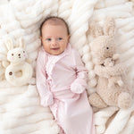 A smiling baby in the Mabel and Honey Amelia Rose Long Sleeve Solid Newborn Gown with Matching Hat, featuring delicate ruffle trim, lies on a soft white blanket surrounded by a plush bunny toy and a bunny-shaped rattle. The gown’s soft Tencel knit ensures cozy comfort for your little one.