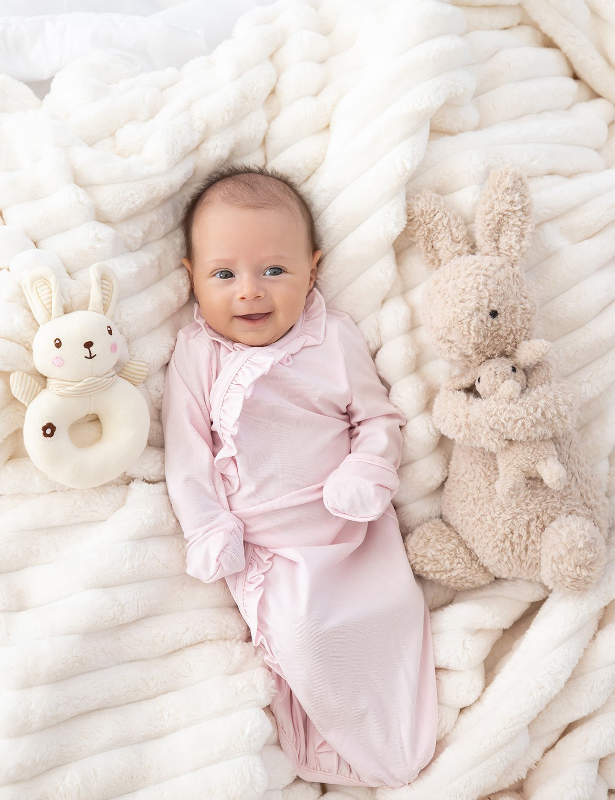 A smiling baby in the Mabel and Honey Amelia Rose Long Sleeve Solid Newborn Gown with Matching Hat, featuring delicate ruffle trim, lies on a soft white blanket surrounded by a plush bunny toy and a bunny-shaped rattle. The gown’s soft Tencel knit ensures cozy comfort for your little one.