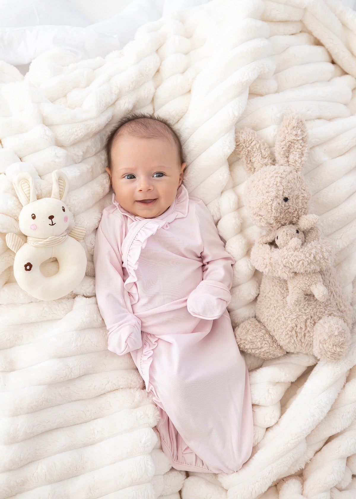 A smiling baby in the Mabel and Honey Amelia Rose Long Sleeve Solid Newborn Gown with Matching Hat, featuring delicate ruffle trim, lies on a soft white blanket surrounded by a plush bunny toy and a bunny-shaped rattle. The gown’s soft Tencel knit ensures cozy comfort for your little one.