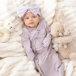 A baby in the Mabel and Honey Soft Meadow Long Sleeve Solid Gown with Matching Hat lies on a soft white blanket, surrounded by a plush bunny and a stuffed rabbit—an adorable scene perfect for capturing that special hospital homecoming outfit.