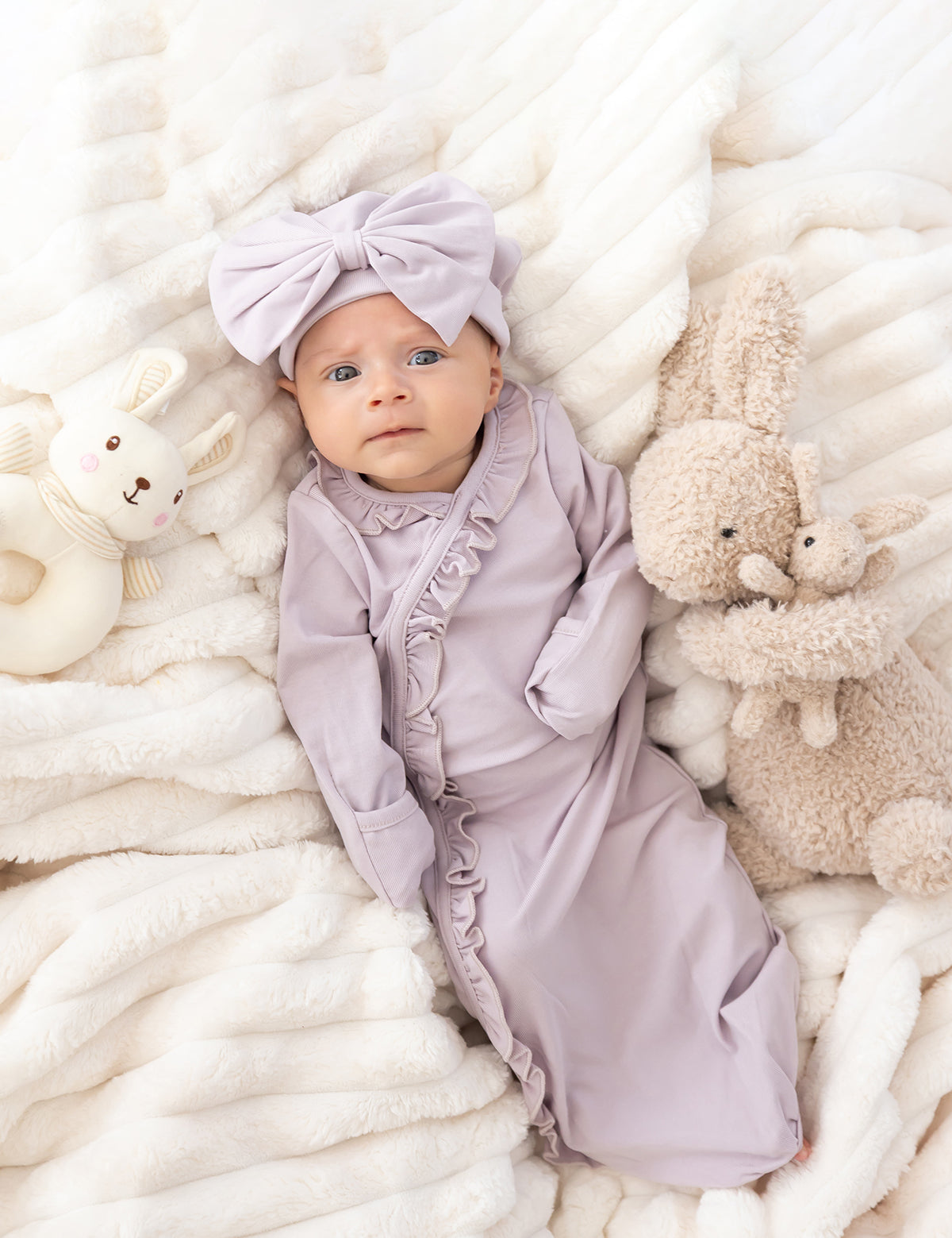 A baby in the Mabel and Honey Soft Meadow Long Sleeve Solid Gown with Matching Hat lies on a soft white blanket, surrounded by a plush bunny and a stuffed rabbit—an adorable scene perfect for capturing that special hospital homecoming outfit.
