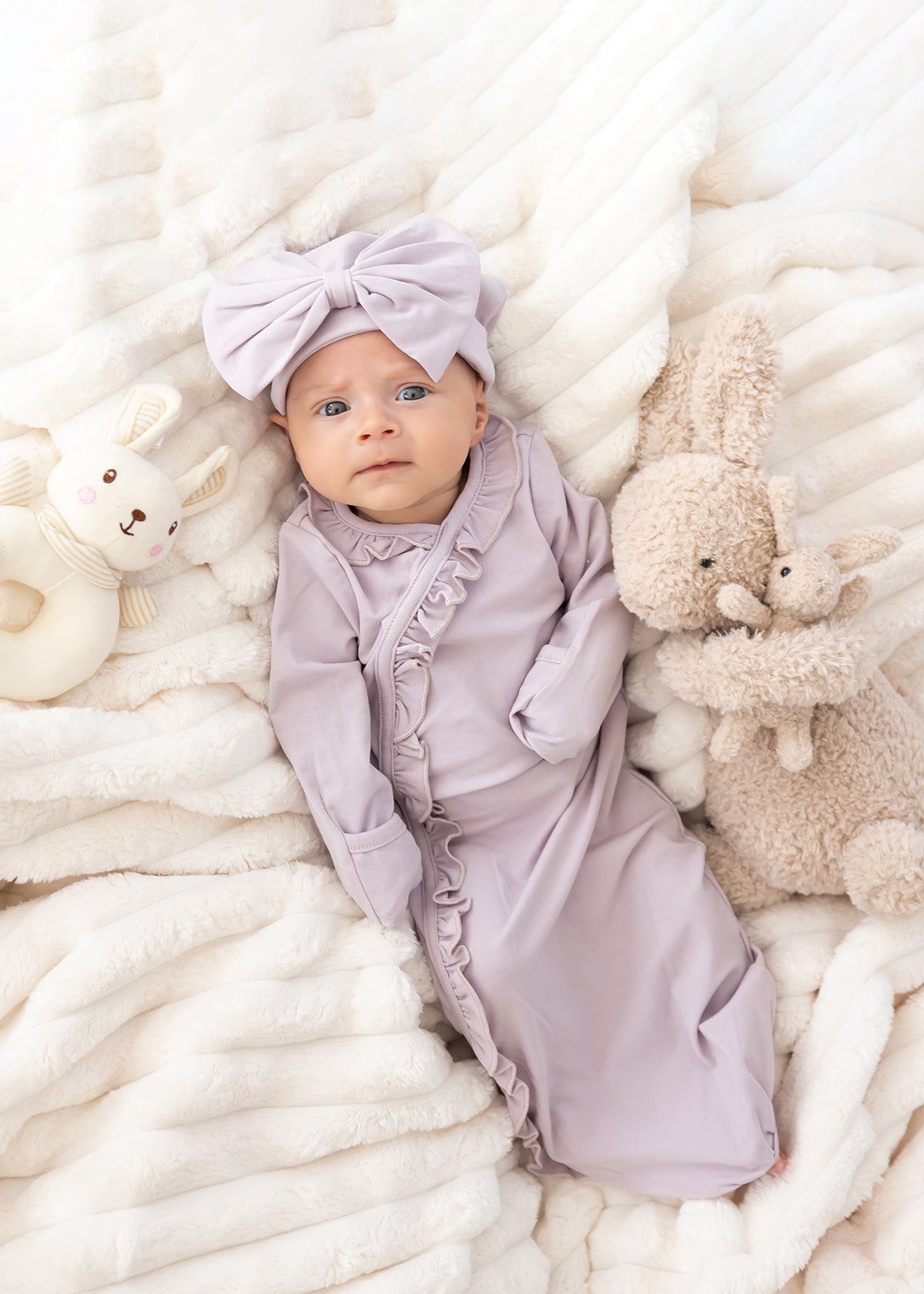 A baby in the Mabel and Honey Soft Meadow Long Sleeve Solid Gown with Matching Hat lies on a soft white blanket, surrounded by a plush bunny and a stuffed rabbit—an adorable scene perfect for capturing that special hospital homecoming outfit.
