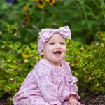 A smiling baby sits barefoot on the ground, surrounded by green plants and yellow flowers, wearing the Mabel and Honey Loretta Long Sleeve Floral Smocked Ruffle Leg Romper with a matching headband.