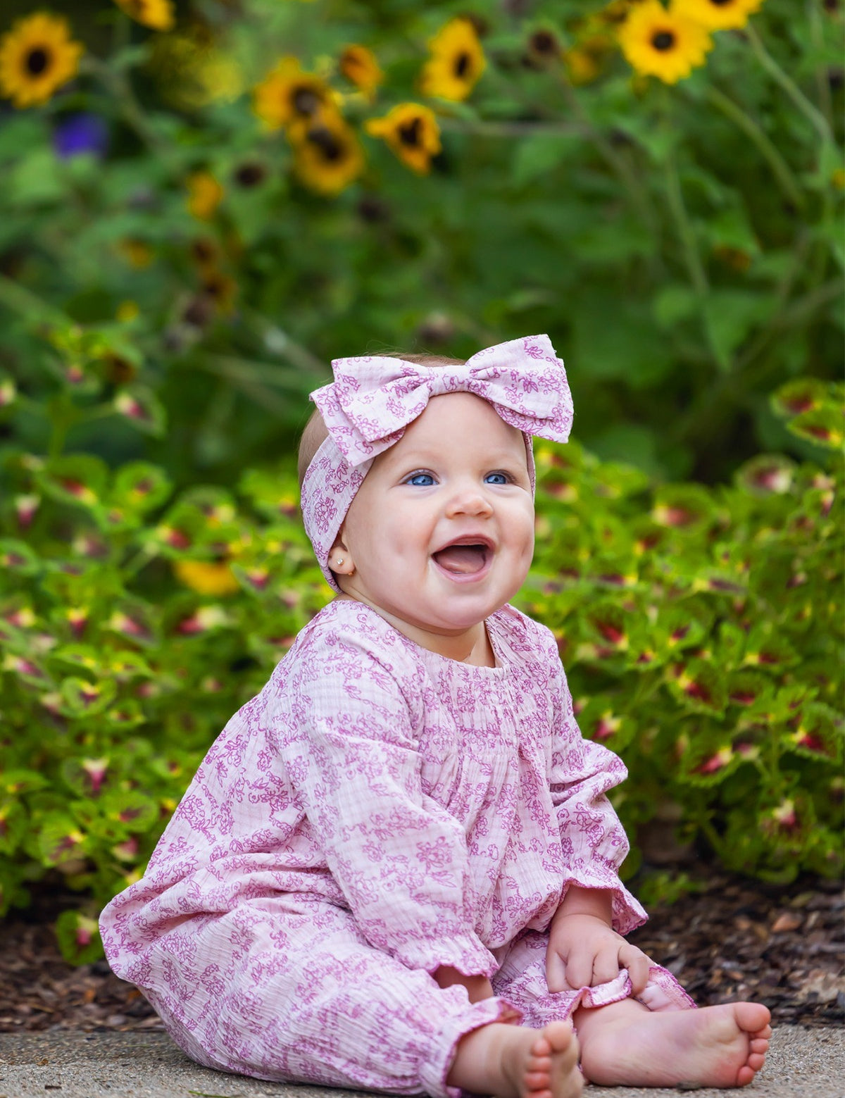 A smiling baby sits barefoot on the ground, surrounded by green plants and yellow flowers, wearing the Mabel and Honey Loretta Long Sleeve Floral Smocked Ruffle Leg Romper with a matching headband.