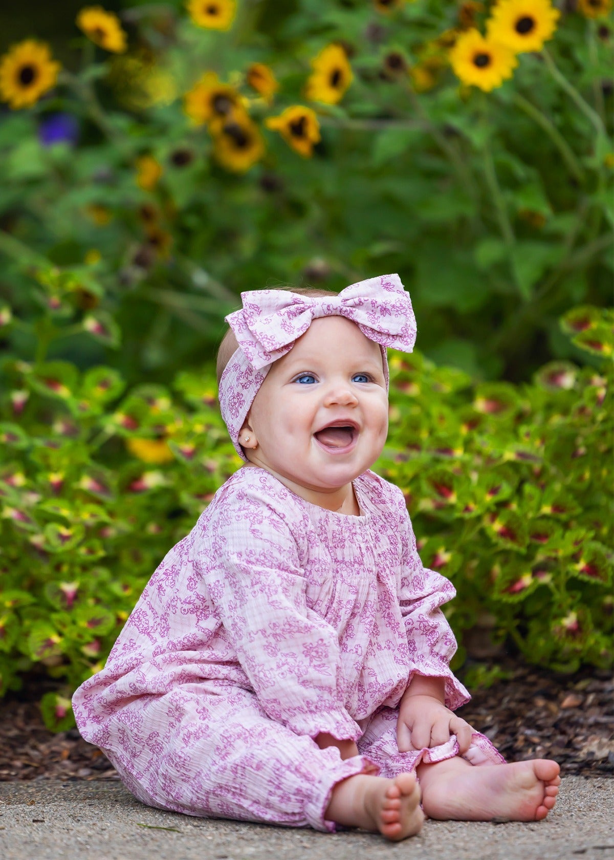 A smiling baby sits barefoot on the ground, surrounded by green plants and yellow flowers, wearing the Mabel and Honey Loretta Long Sleeve Floral Smocked Ruffle Leg Romper with a matching headband.