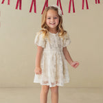 A smiling young girl stands indoors wearing the Isobella and Chloe Golden Goose Short Sleeve Floral Sequin Ruffle Sleeve Empire Waist Dress. Red ribbon bows hang on the wall behind her. She pairs the dress with light blue shoes and a matching white bow in her long blonde hair, making it a perfect choice for a holiday party.