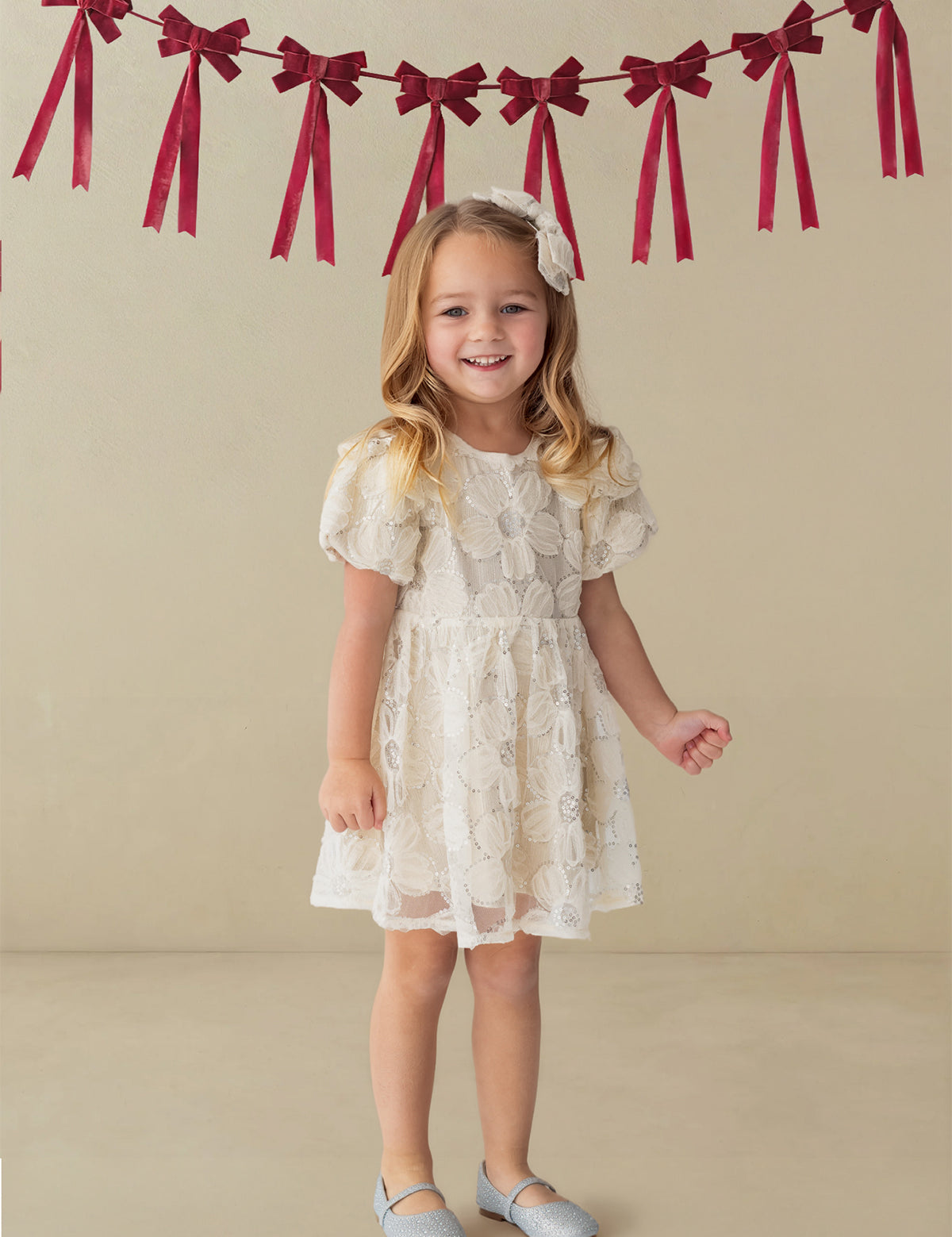 A smiling young girl stands indoors wearing the Isobella and Chloe Golden Goose Short Sleeve Floral Sequin Ruffle Sleeve Empire Waist Dress. Red ribbon bows hang on the wall behind her. She pairs the dress with light blue shoes and a matching white bow in her long blonde hair, making it a perfect choice for a holiday party.
