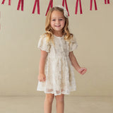 A smiling young girl stands indoors wearing the Isobella and Chloe Golden Goose Short Sleeve Floral Sequin Ruffle Sleeve Empire Waist Dress. Red ribbon bows hang on the wall behind her. She pairs the dress with light blue shoes and a matching white bow in her long blonde hair, making it a perfect choice for a holiday party.