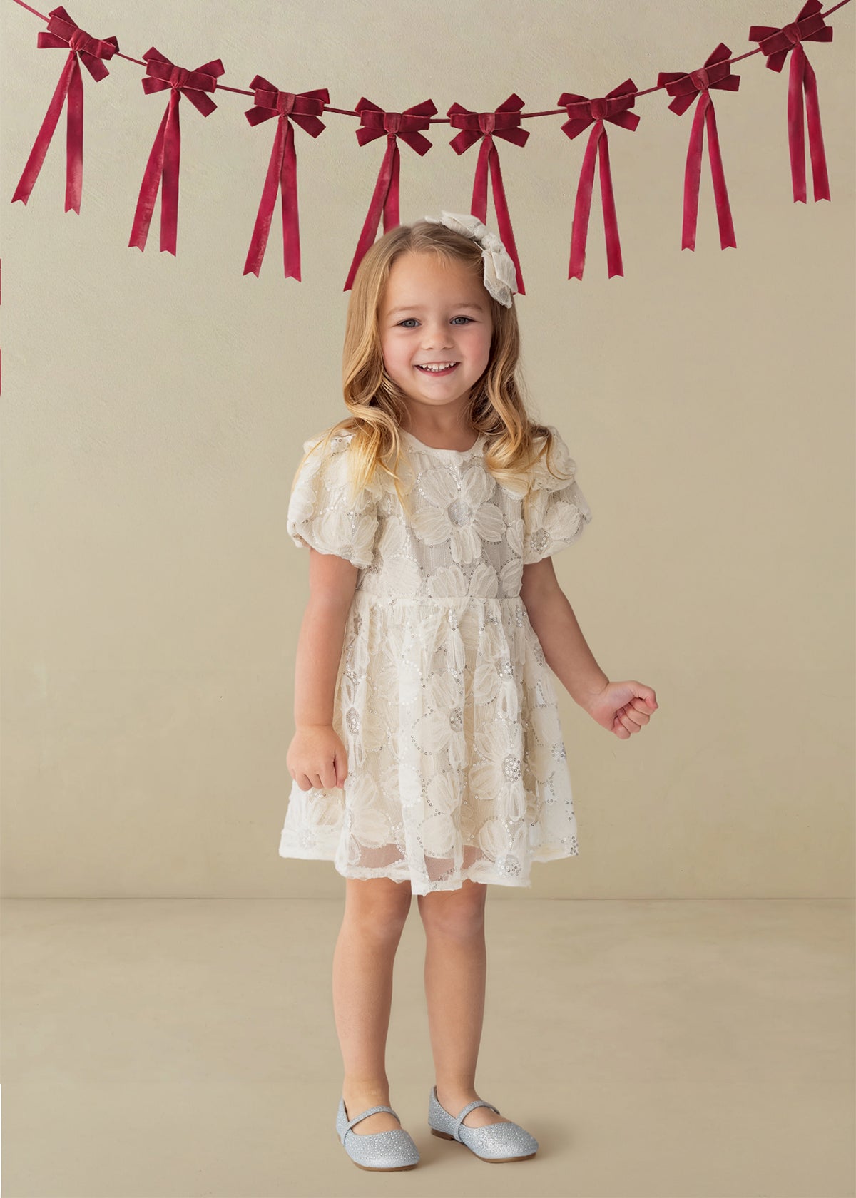 A smiling young girl stands indoors wearing the Isobella and Chloe Golden Goose Short Sleeve Floral Sequin Ruffle Sleeve Empire Waist Dress. Red ribbon bows hang on the wall behind her. She pairs the dress with light blue shoes and a matching white bow in her long blonde hair, making it a perfect choice for a holiday party.