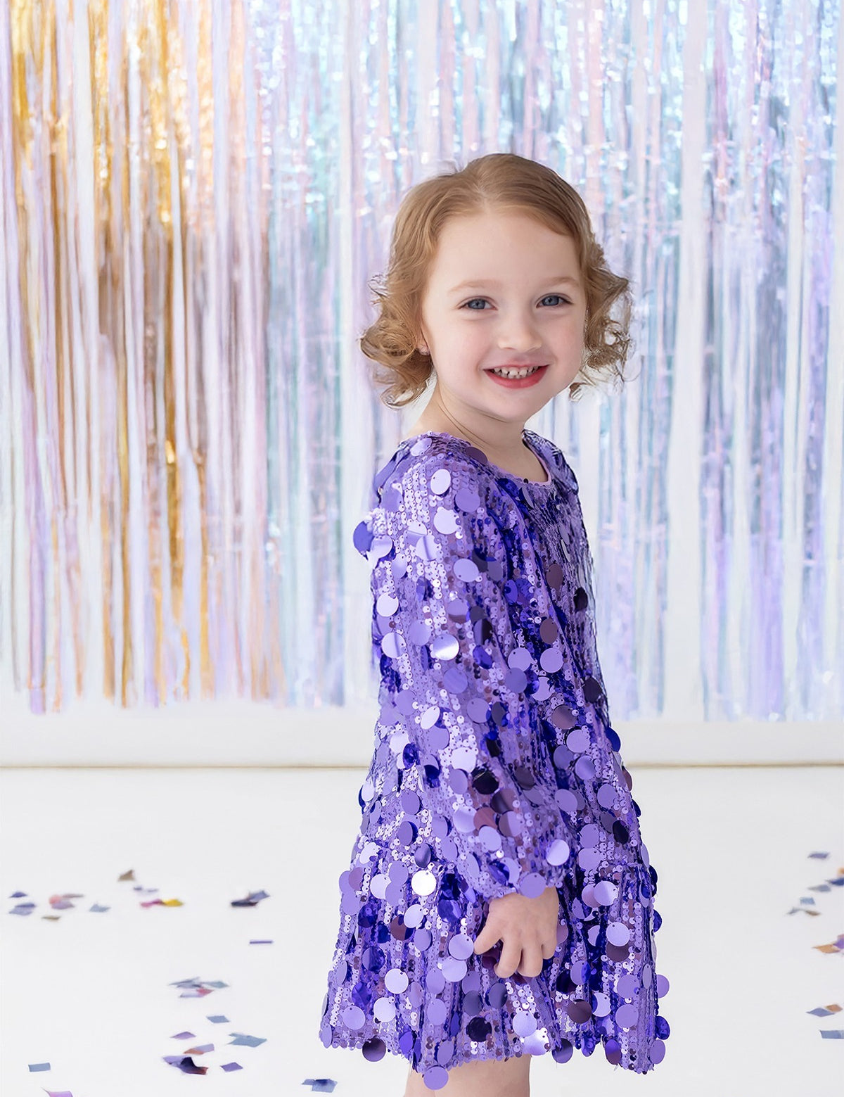 A young child wearing the Isobella and Chloe “Shake it Off” Bishop Sleeve Sequin Ruffle Hem Mini Dress smiles while standing in front of shiny pastel streamers, with confetti scattered on the white floor.