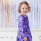 A young child wearing the Isobella and Chloe “Shake it Off” Bishop Sleeve Sequin Ruffle Hem Mini Dress smiles while standing in front of shiny pastel streamers, with confetti scattered on the white floor.